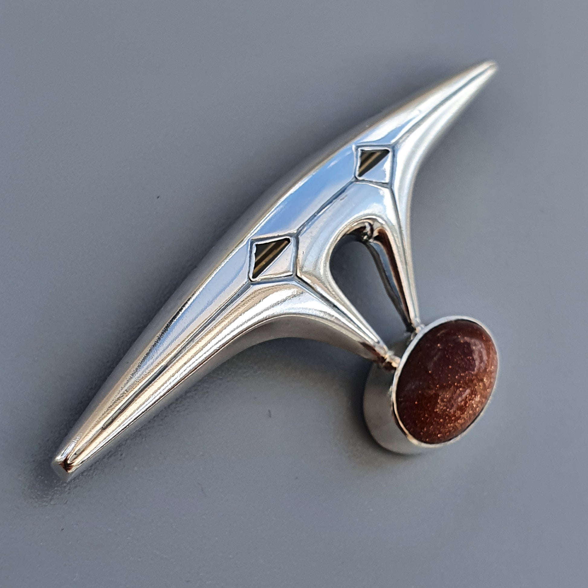 Futuristic silver brooch or pin with a sleek, angular design and a reddish-brown gemstone accent.