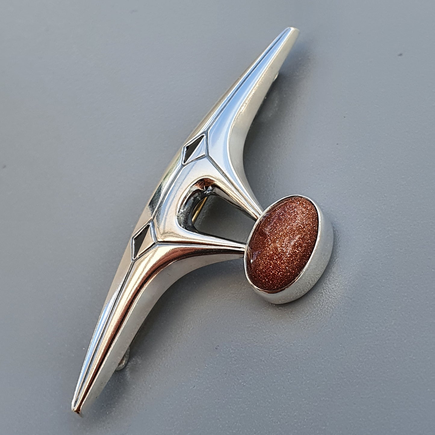 Shiny metallic car hood ornament with a sleek, angular design and a reddish-brown oval accent.