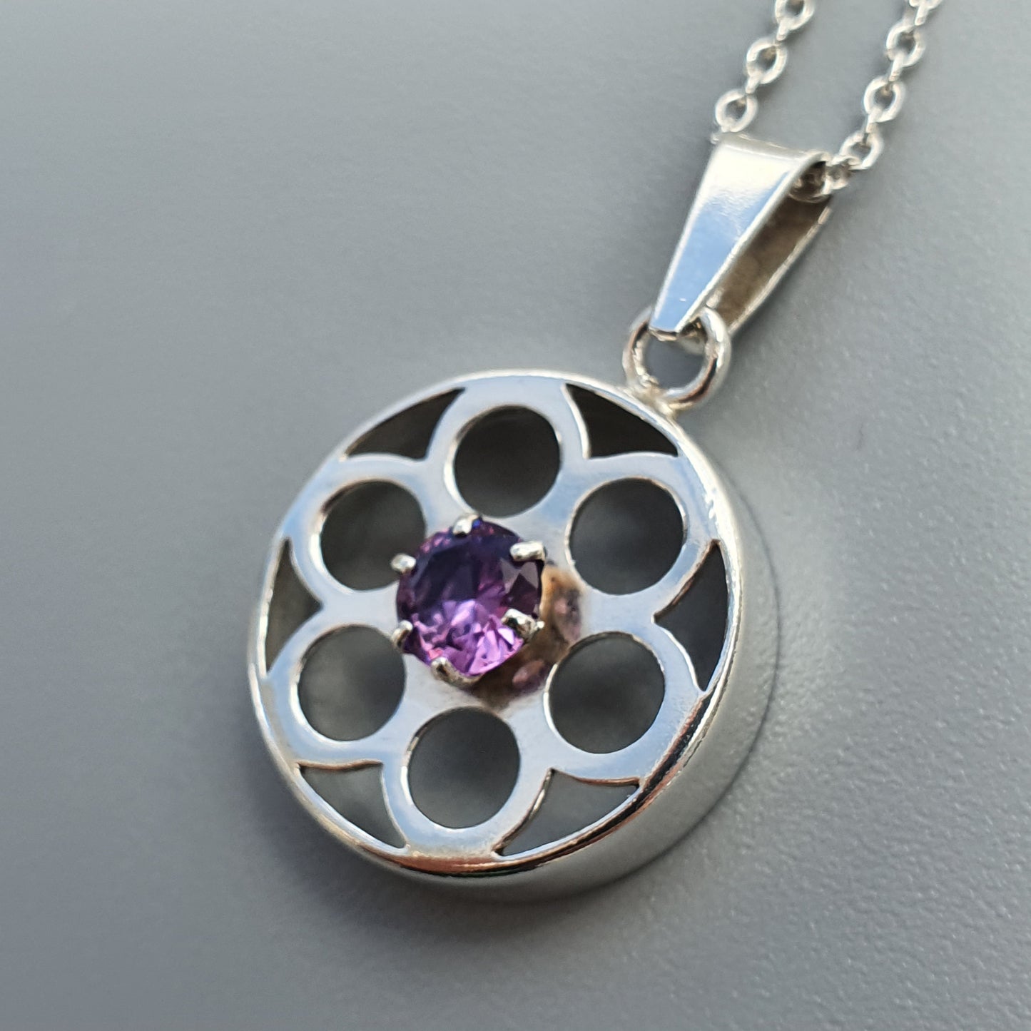 Silver pendant necklace featuring a circular design with flower-like cutouts and a central purple gemstone.