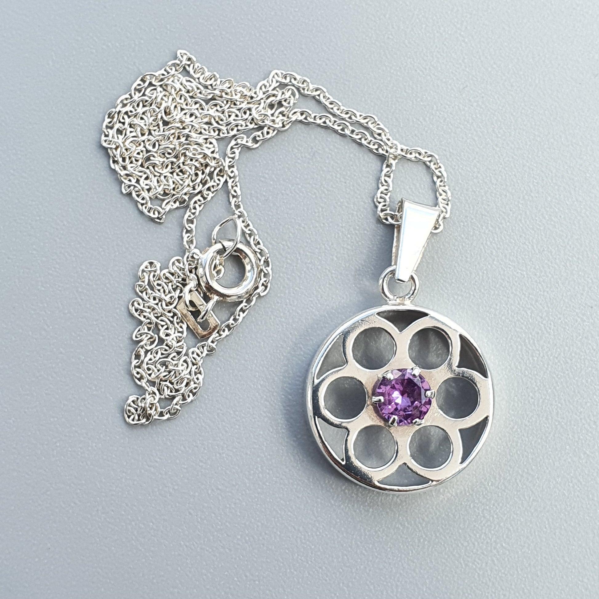 Silver pendant necklace with a circular flower-shaped charm featuring a purple gemstone in the center.