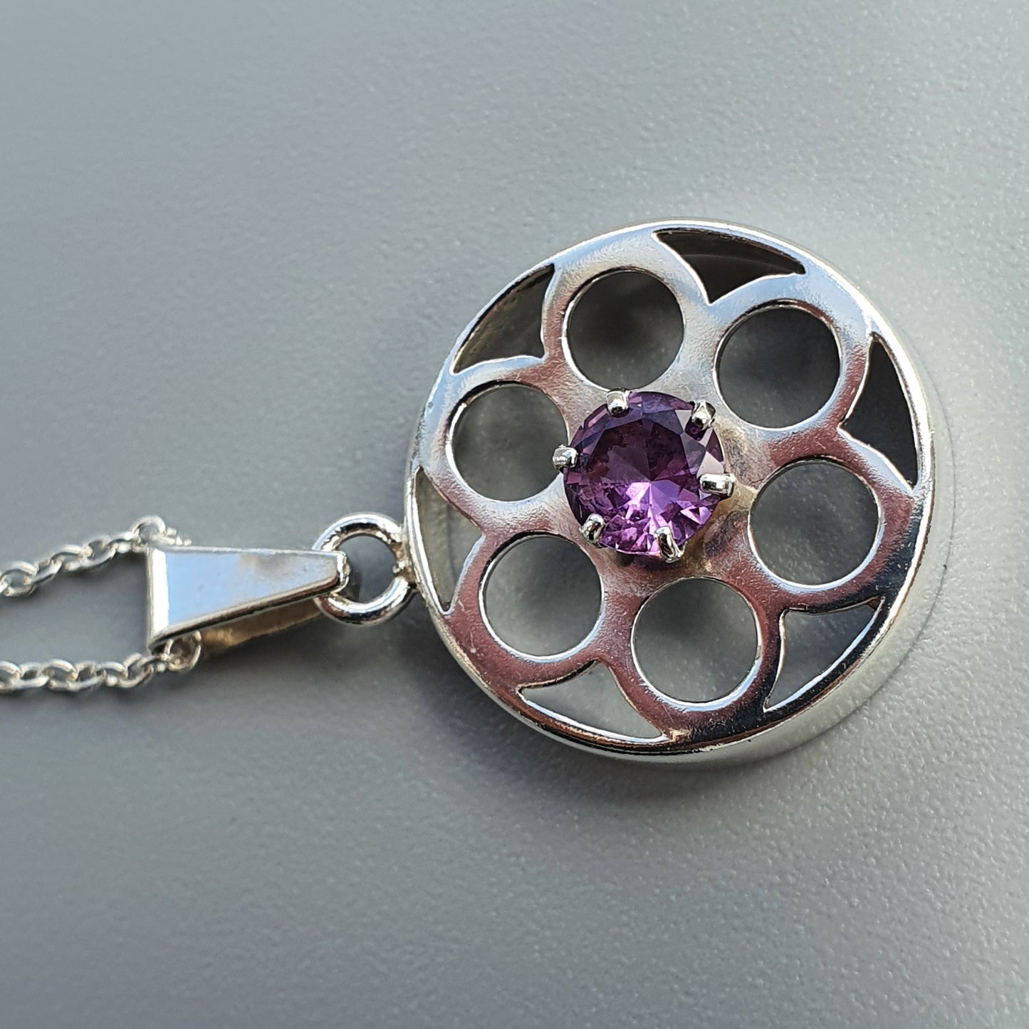 Circular silver pendant with a purple gemstone center and multiple circular cutouts.