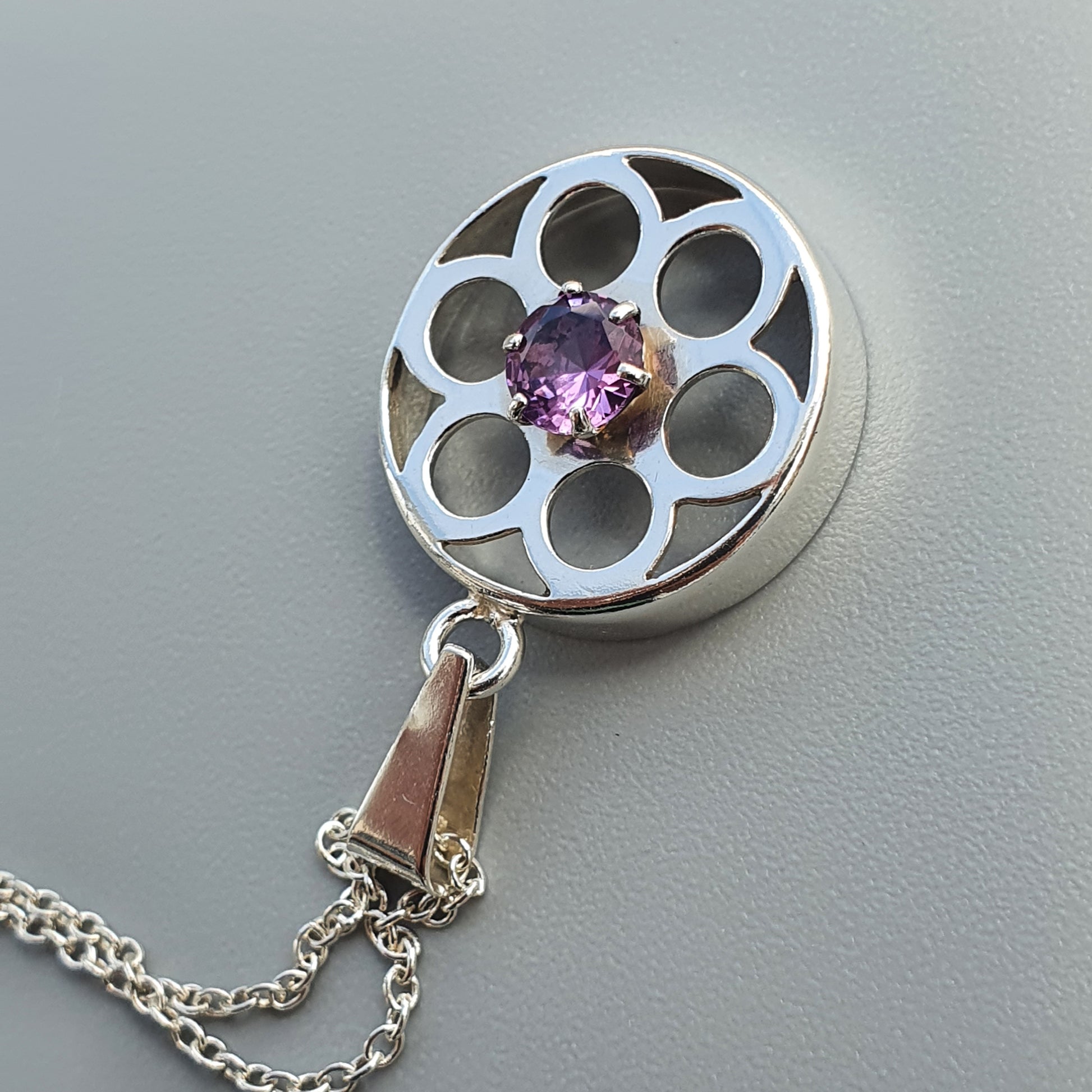 Circular silver pendant with a flower-like design and a purple gemstone at its center, attached to a chain.