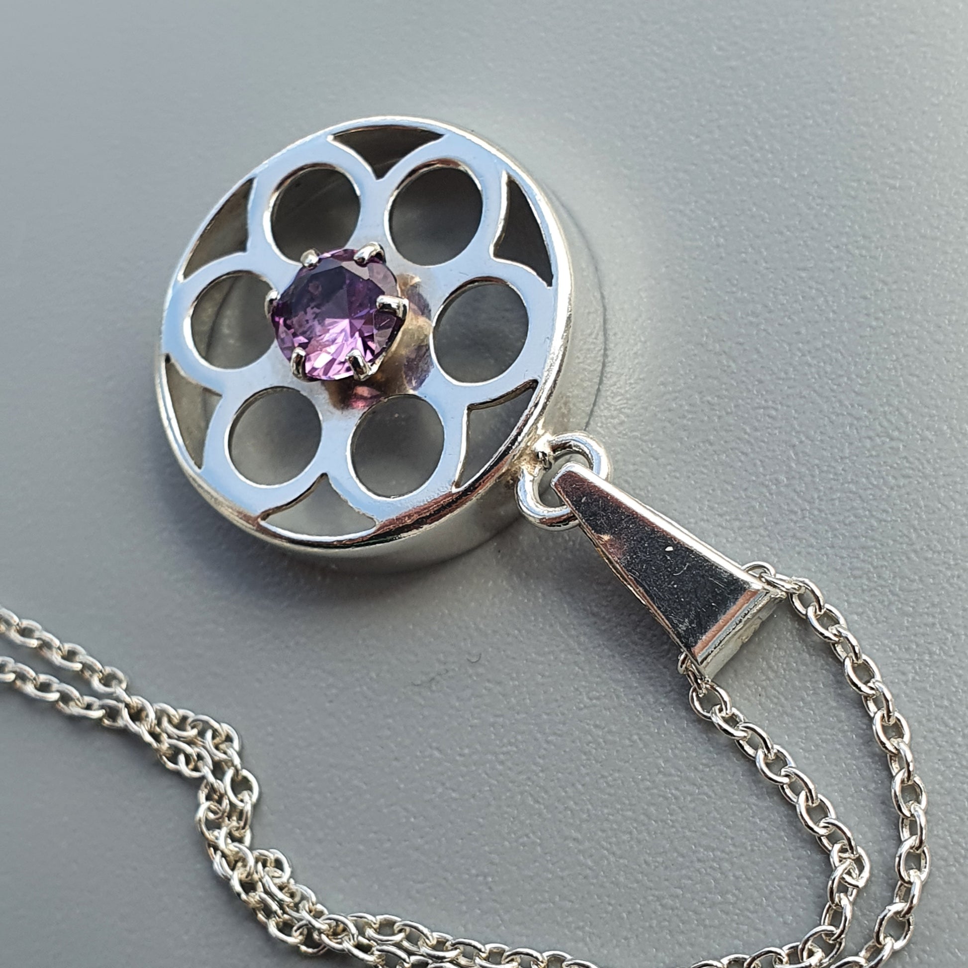 Silver pendant with a circular floral design and purple gemstone center, attached to a chain.