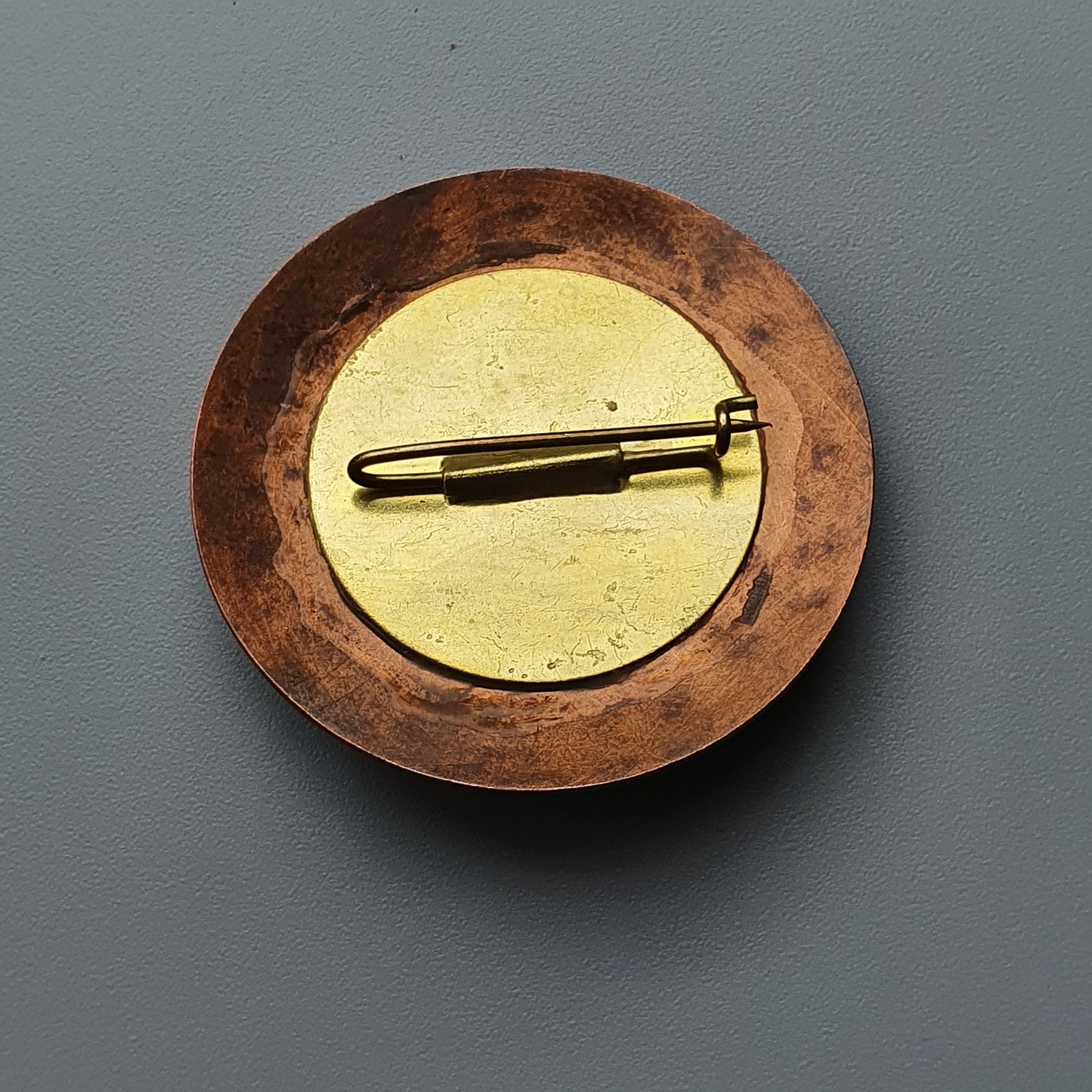 Circular brooch with a copper-colored outer ring and gold-toned center featuring a pen-shaped pin.