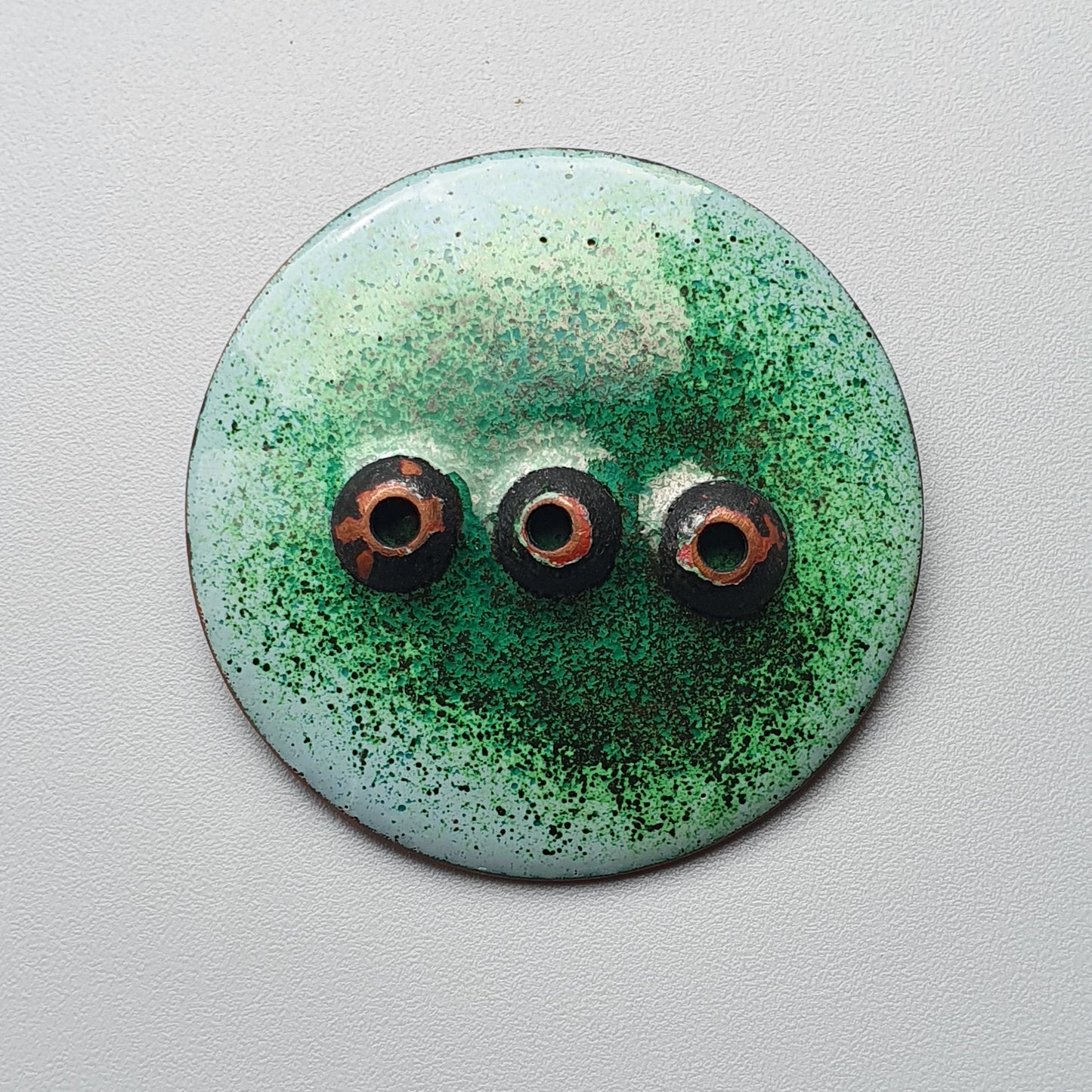 Circular green button or brooch with three eye-like protrusions.