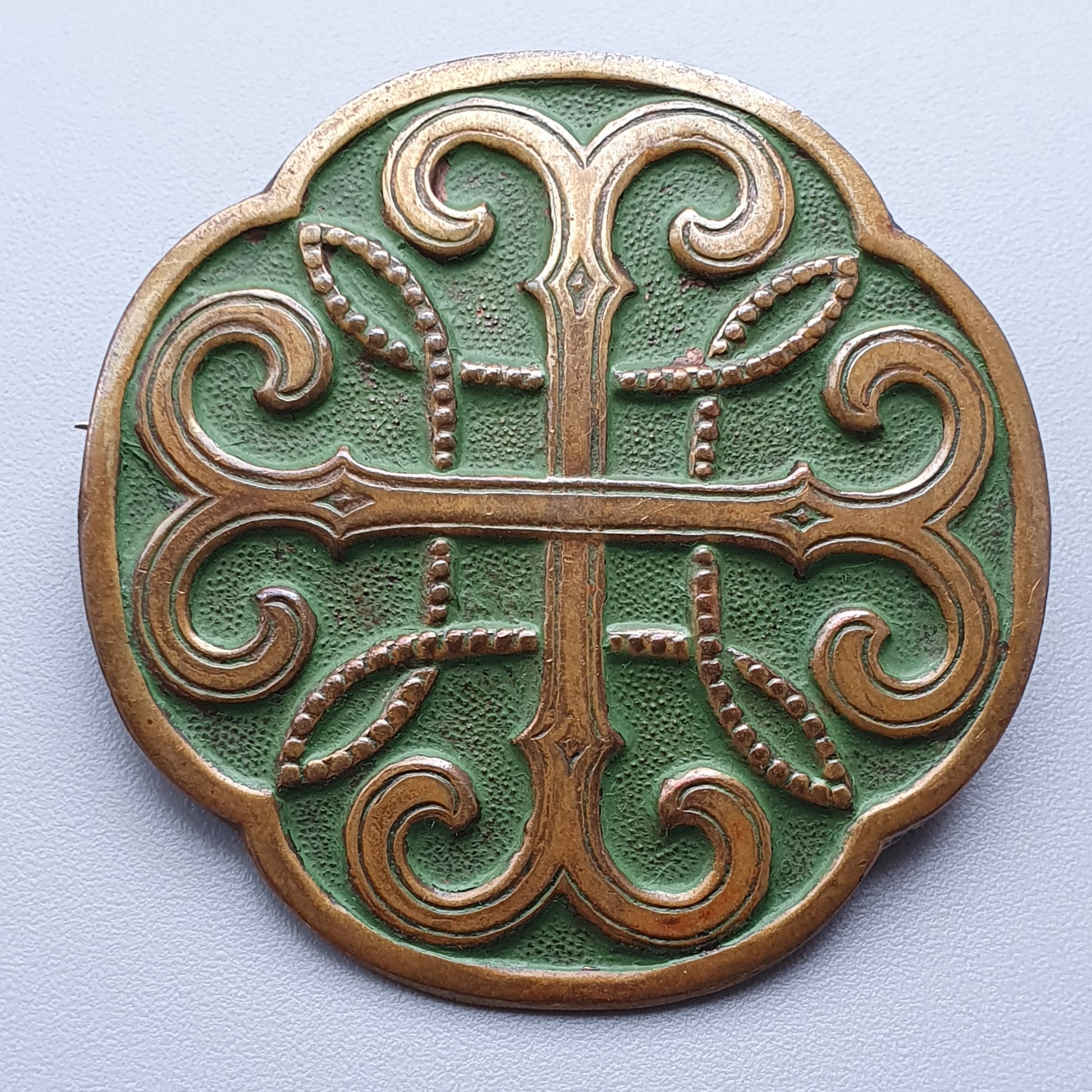 Ornate circular medallion with a symmetrical cross-like design in bronze relief on a green background.