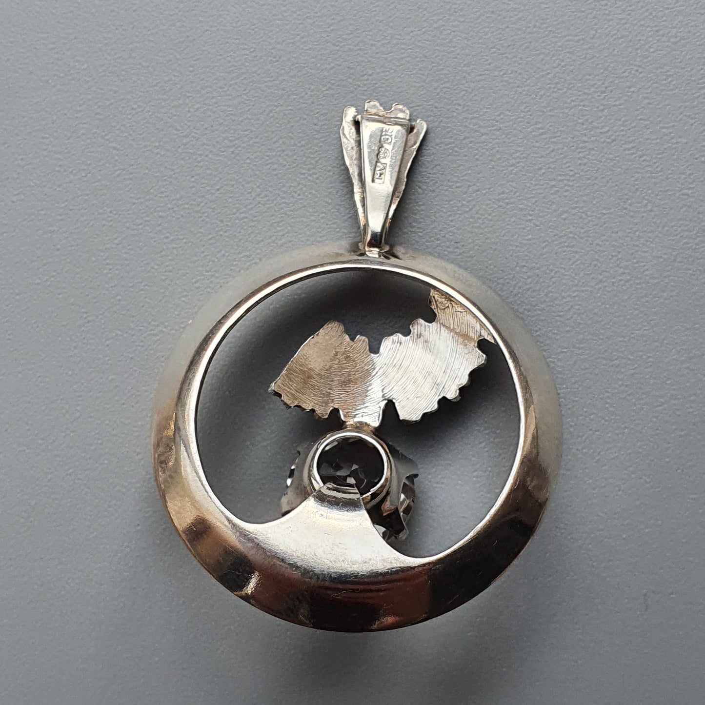 Silver pendant with a circular frame containing metallic leaf-like shapes and a small gemstone.