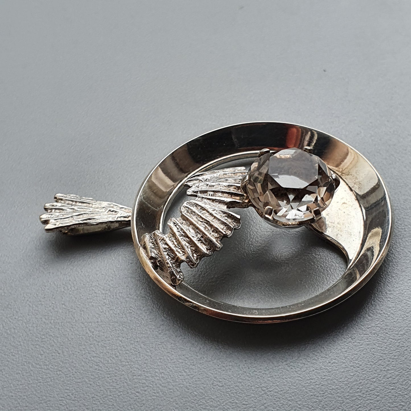 Circular silver pendant featuring a wing design and a clear gemstone.