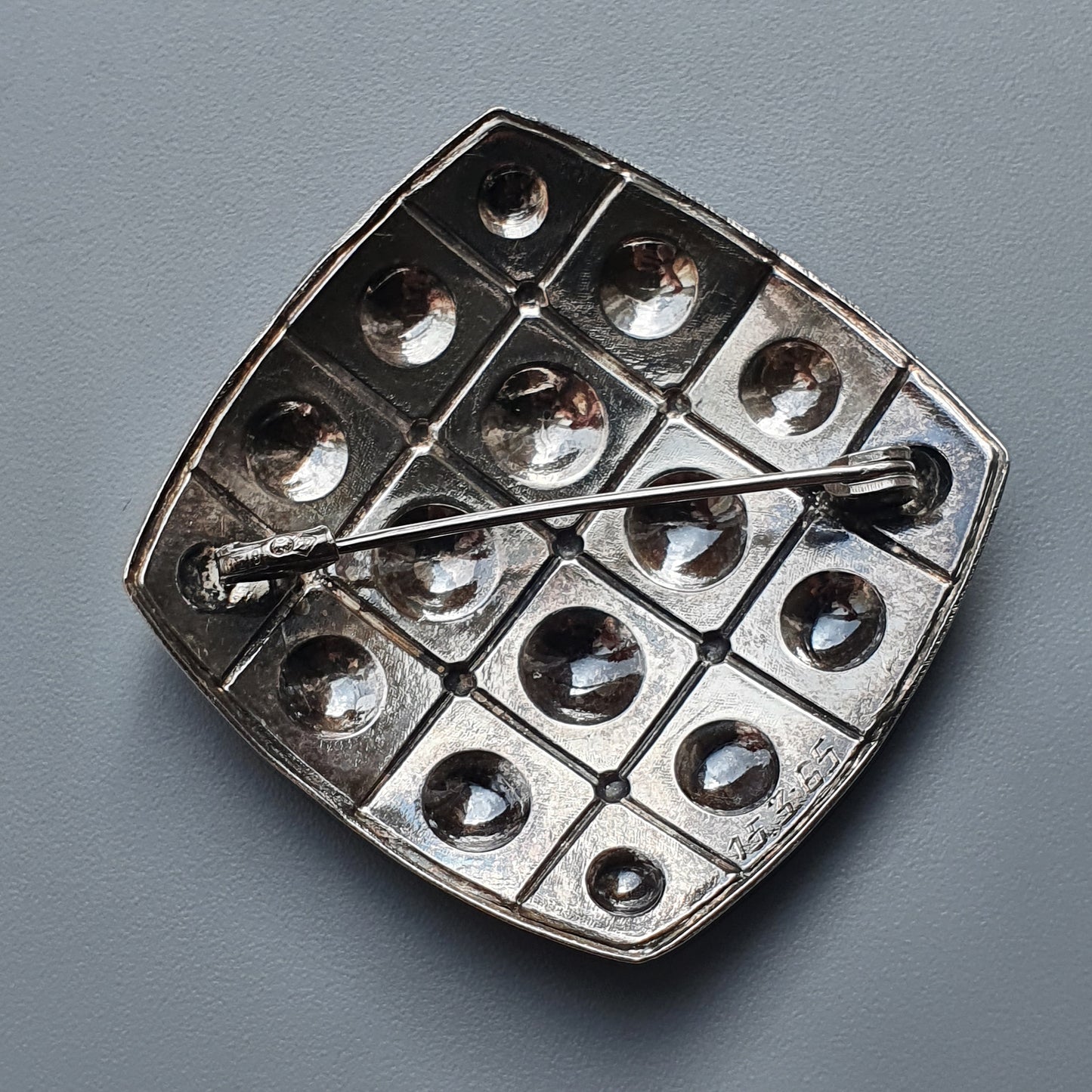 Metallic brooch or pin with a geometric grid pattern and circular indentations.