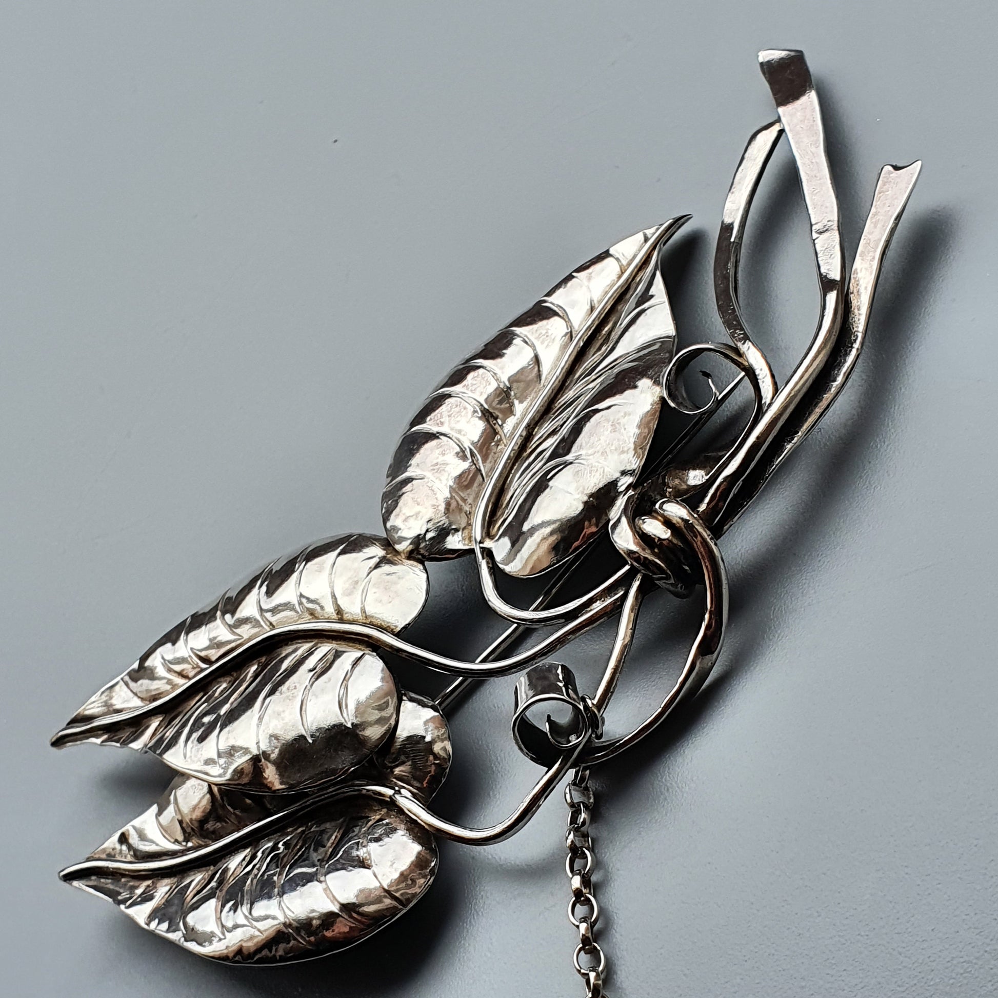 Ornate silver brooch shaped like leaves or foliage with a dangling chain.