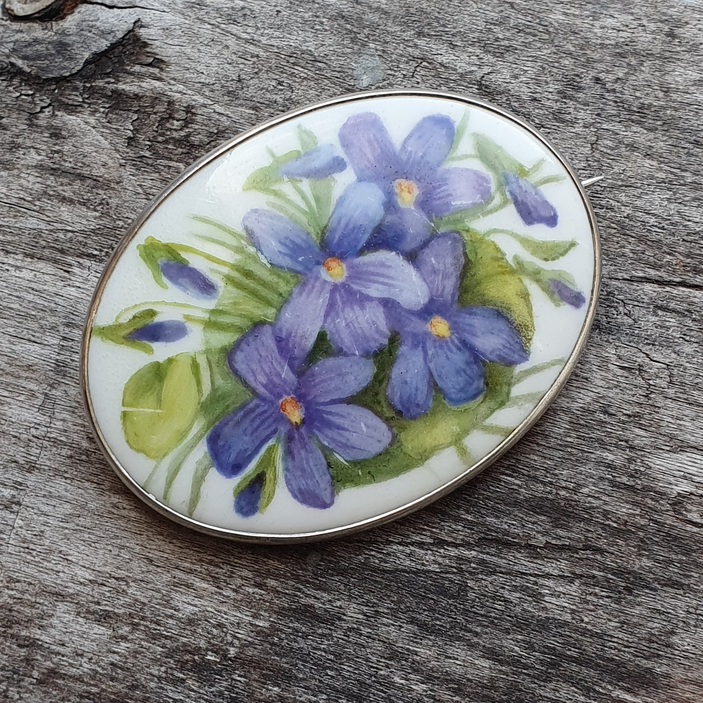 Oval brooch or pendant featuring painted purple flowers on a white background.