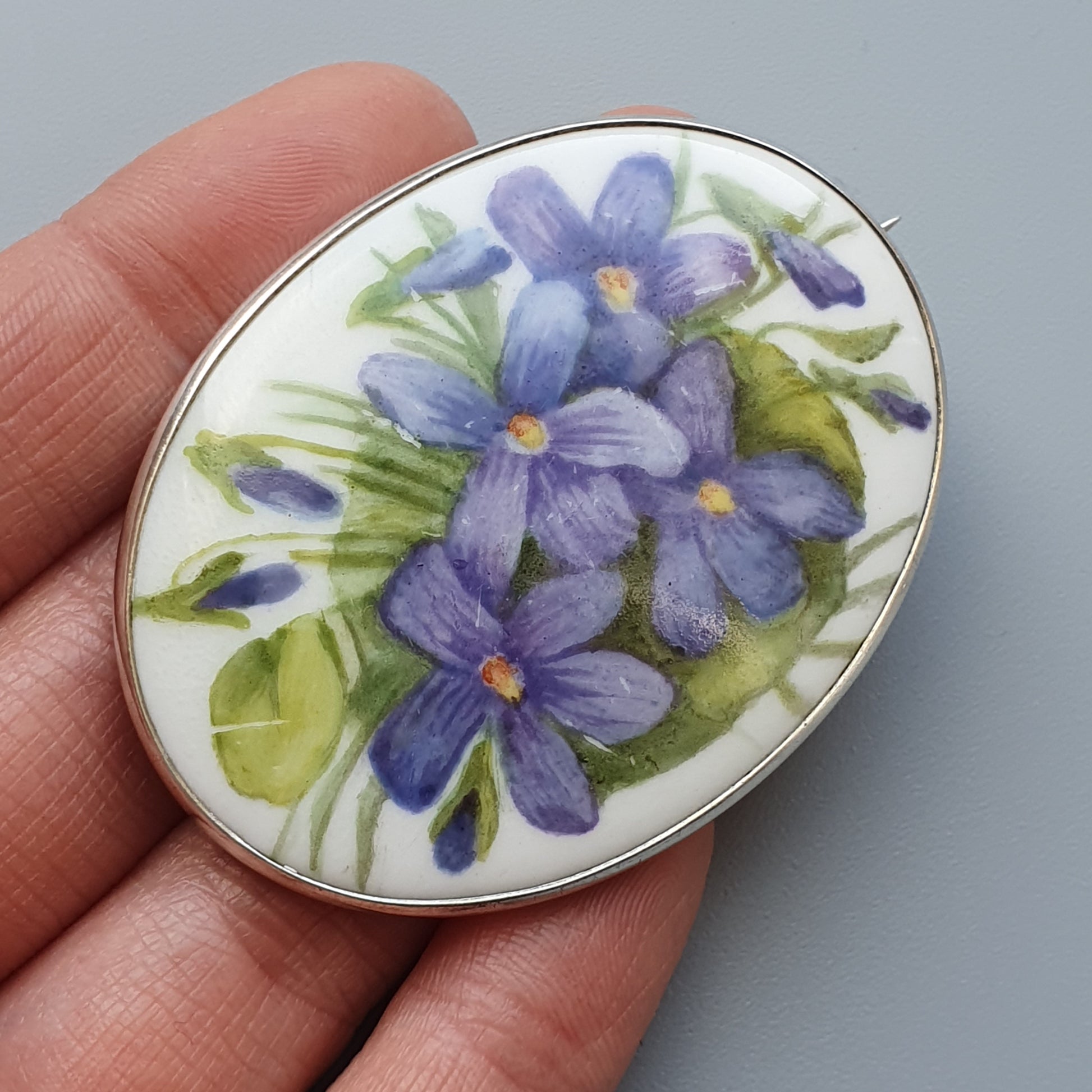 Oval brooch featuring painted purple violets on a white background.