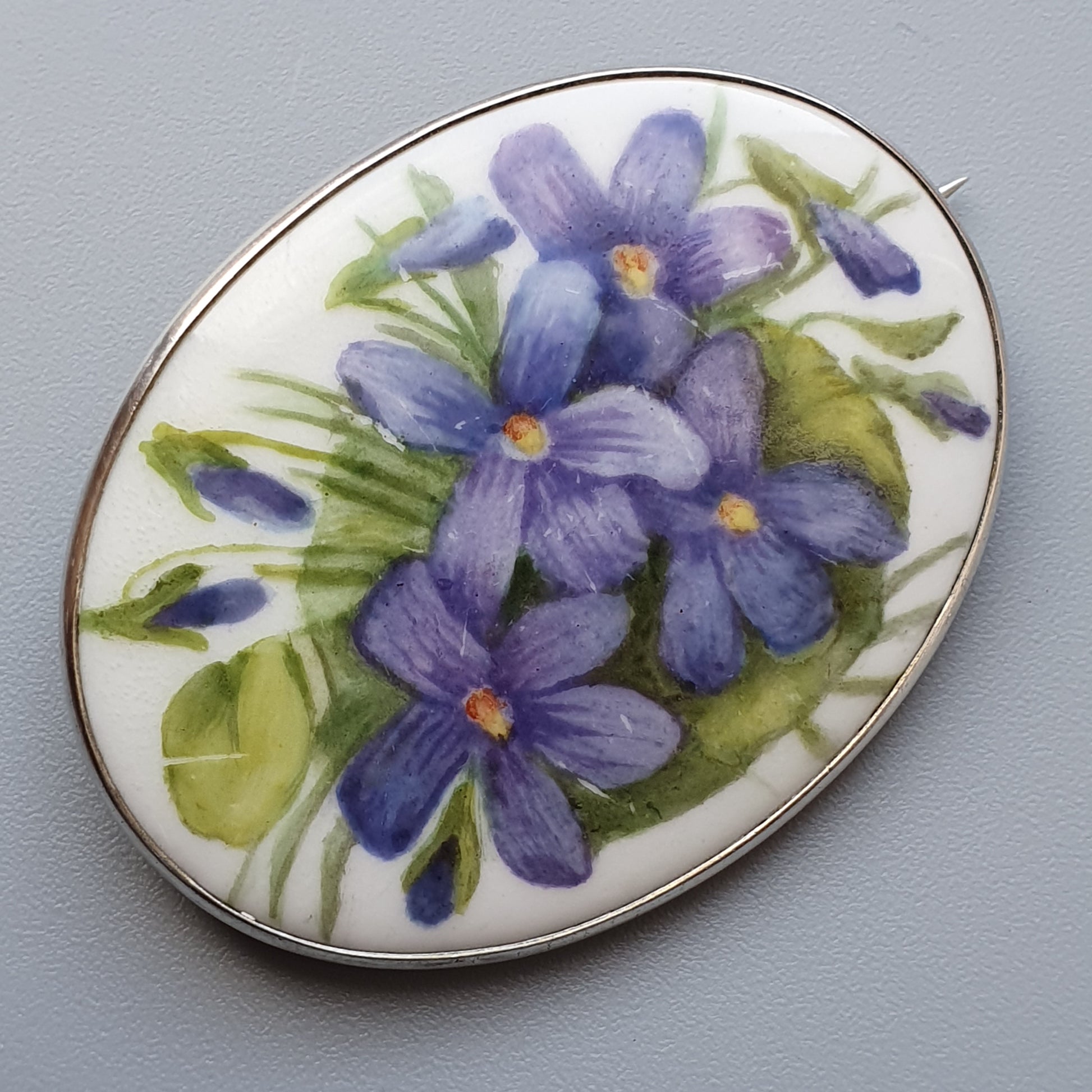 Oval brooch featuring hand-painted purple violets on a porcelain or enamel surface with a metallic border.