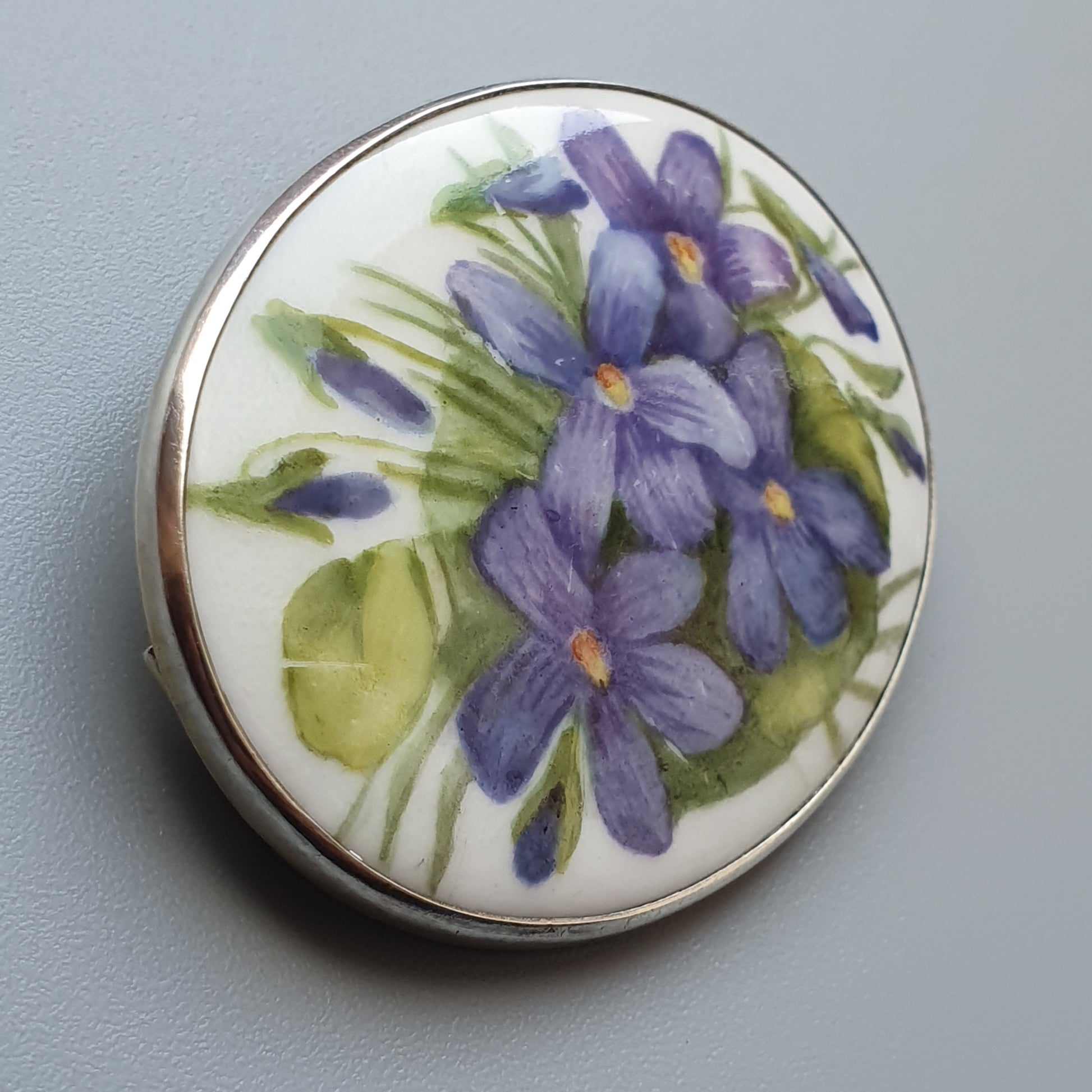 Decorative porcelain brooch featuring painted purple violets with green leaves.