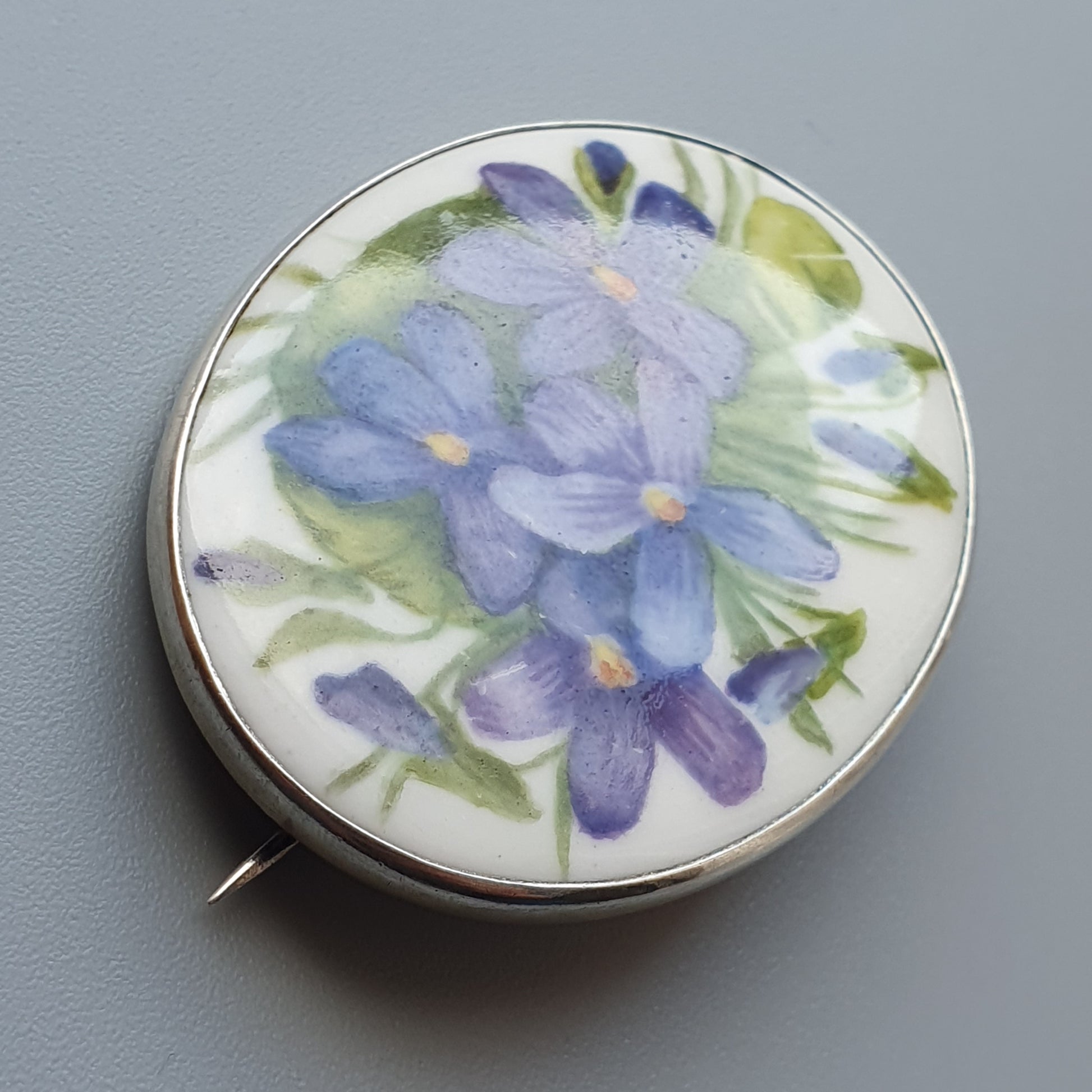 Circular porcelain brooch featuring painted purple flowers with green leaves.