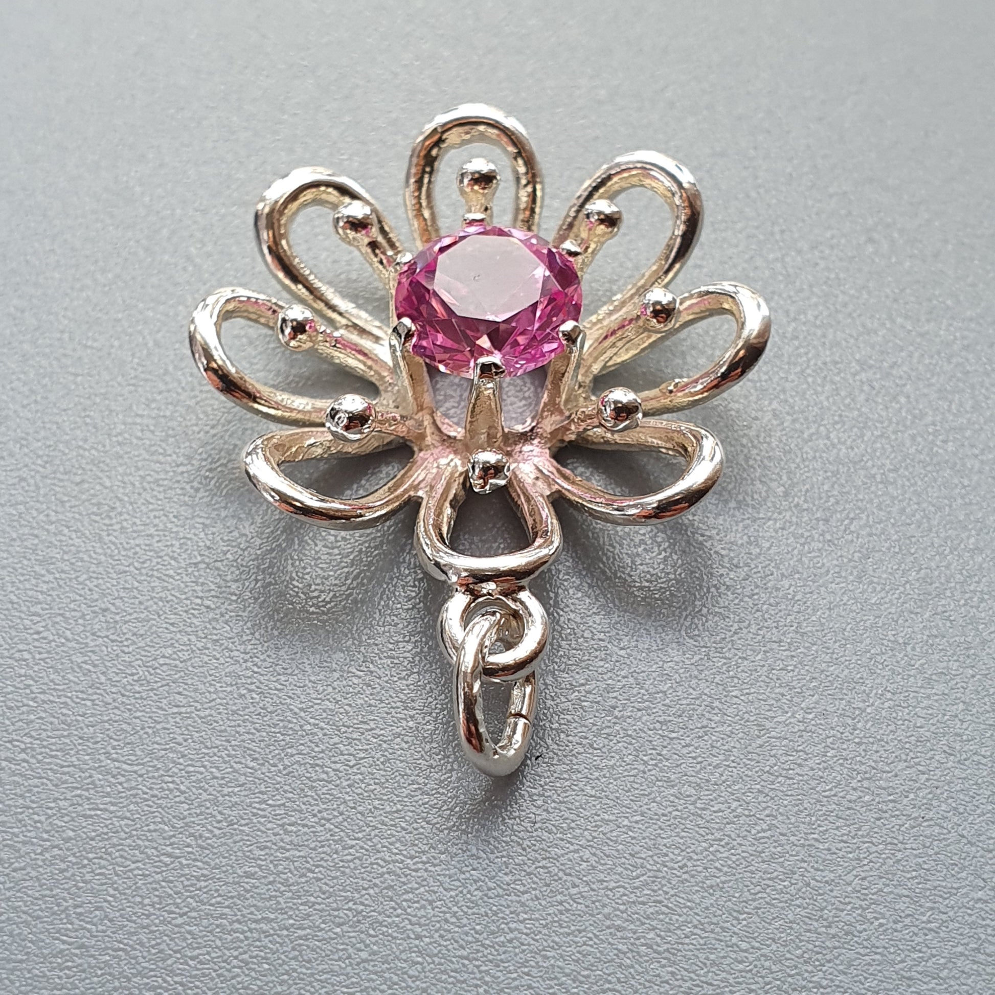 Decorative gold-toned charm or pendant shaped like a stylized flower with a pink gemstone center.