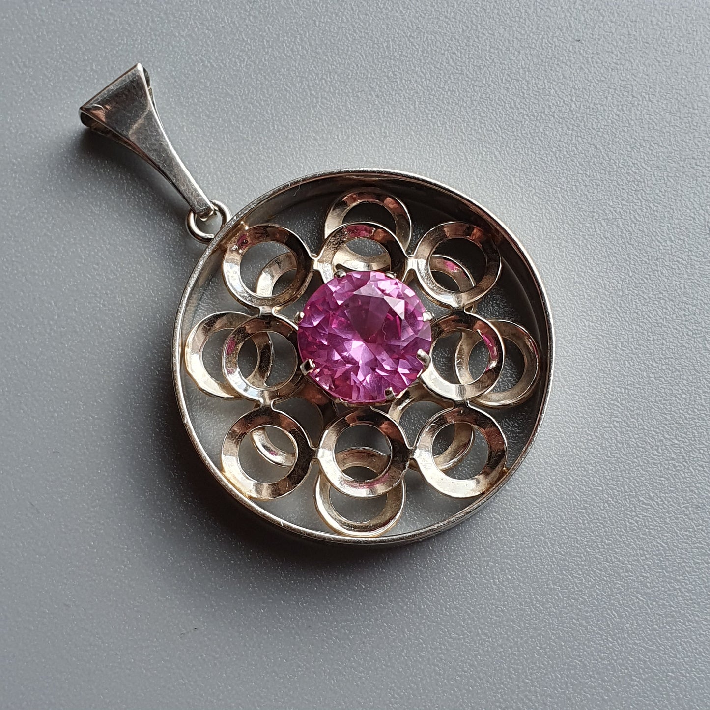 Circular silver pendant with a pink gemstone center surrounded by a floral-like design.