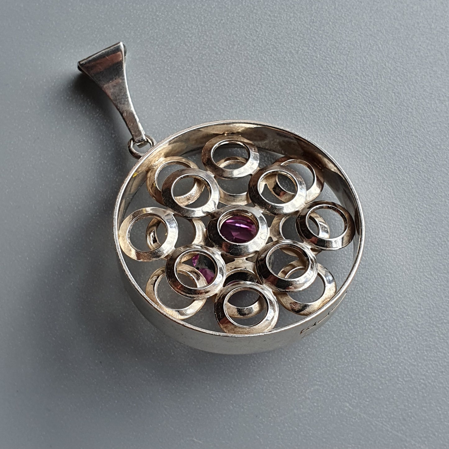 Circular silver pendant with a pattern of loops and a central purple gemstone.