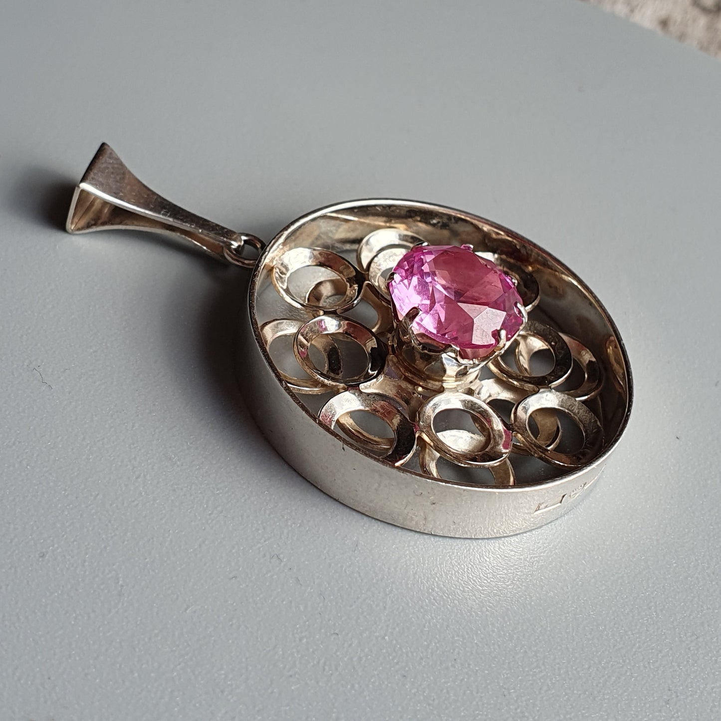 Teardrop-shaped silver pendant with a pink gemstone and decorative swirls.