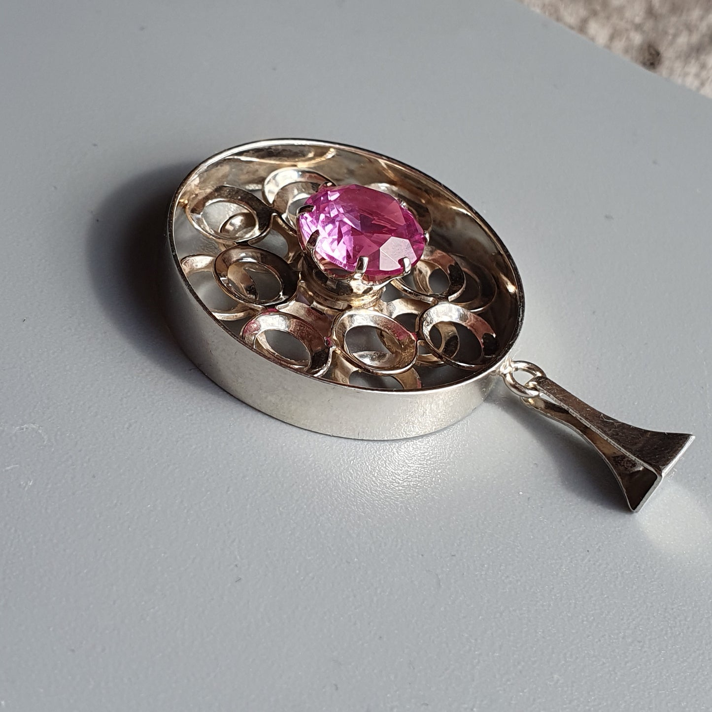 Silver pendant with an oval shape featuring a pink gemstone surrounded by decorative cutouts.