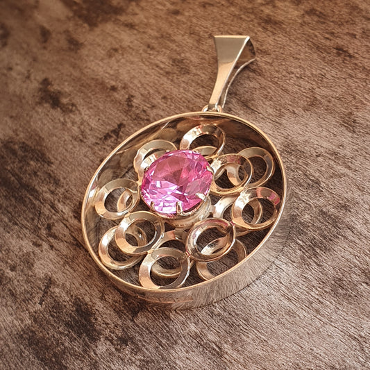 Gold pendant featuring a pink gemstone surrounded by swirling metalwork.