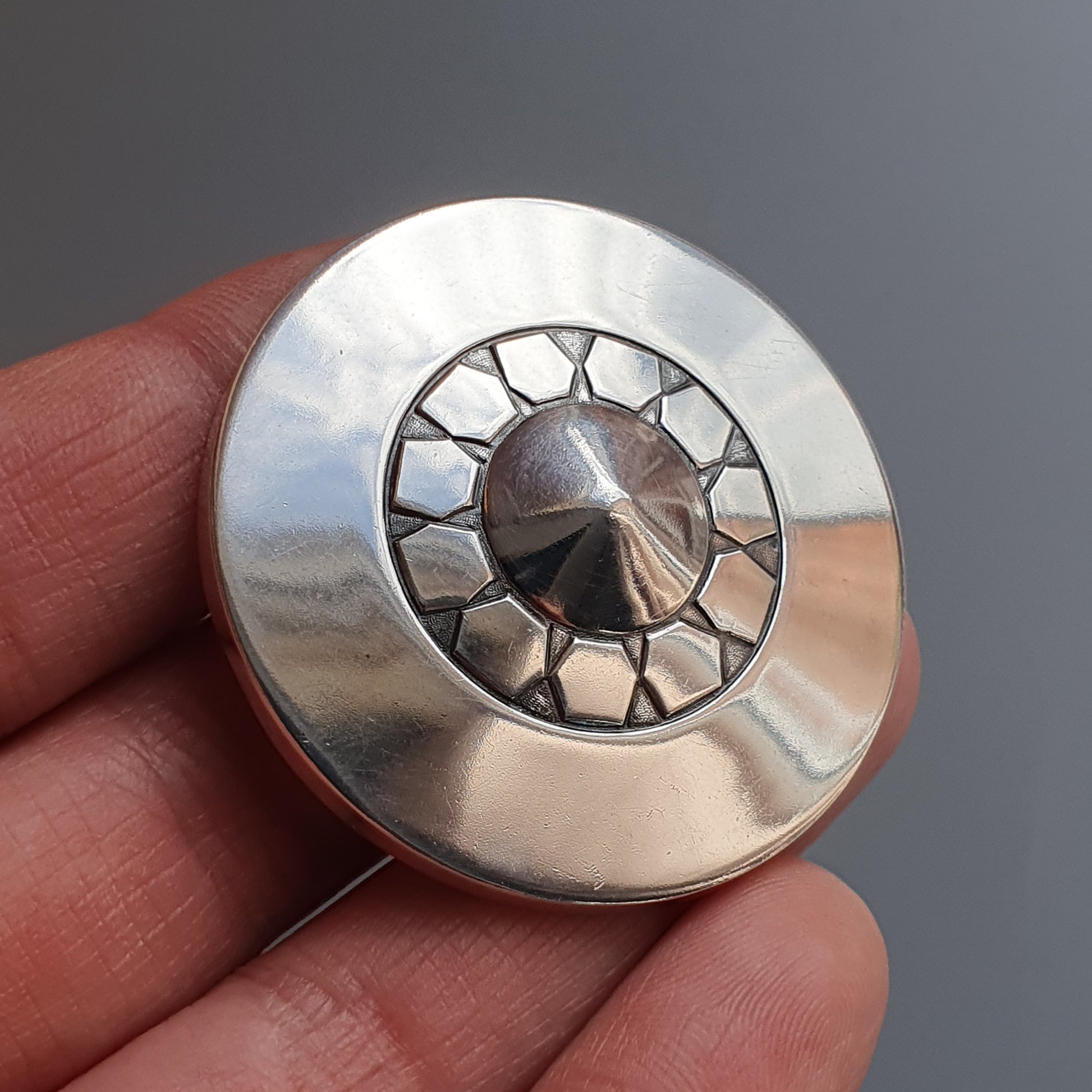 Circular silver pendant or brooch with a geometric sunburst design in the center.