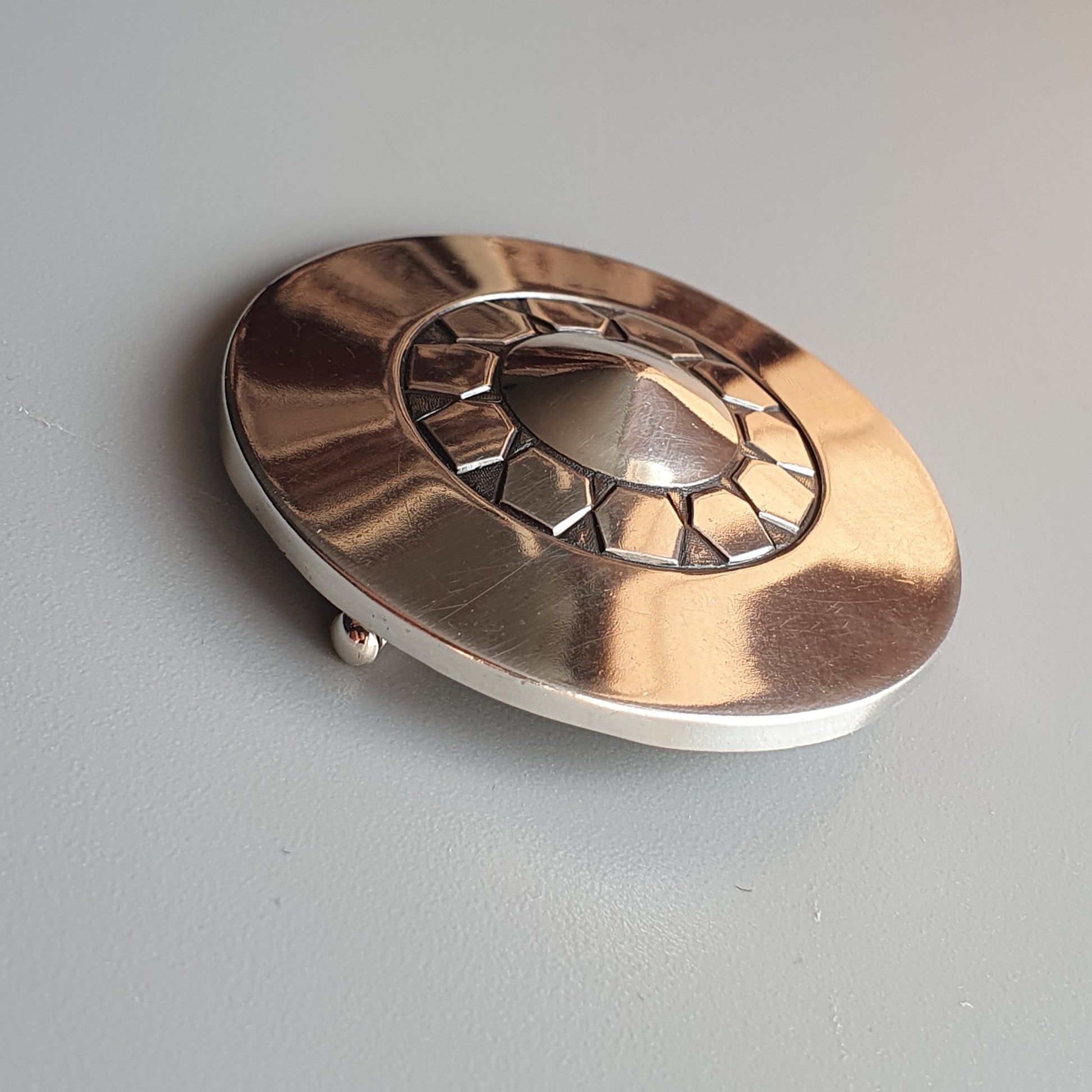 Metallic oval brooch or pin with a geometric sunburst design in the center.