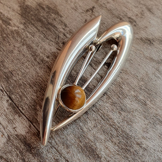 Silver leaf-shaped brooch with a round tiger’s eye gemstone at its center.