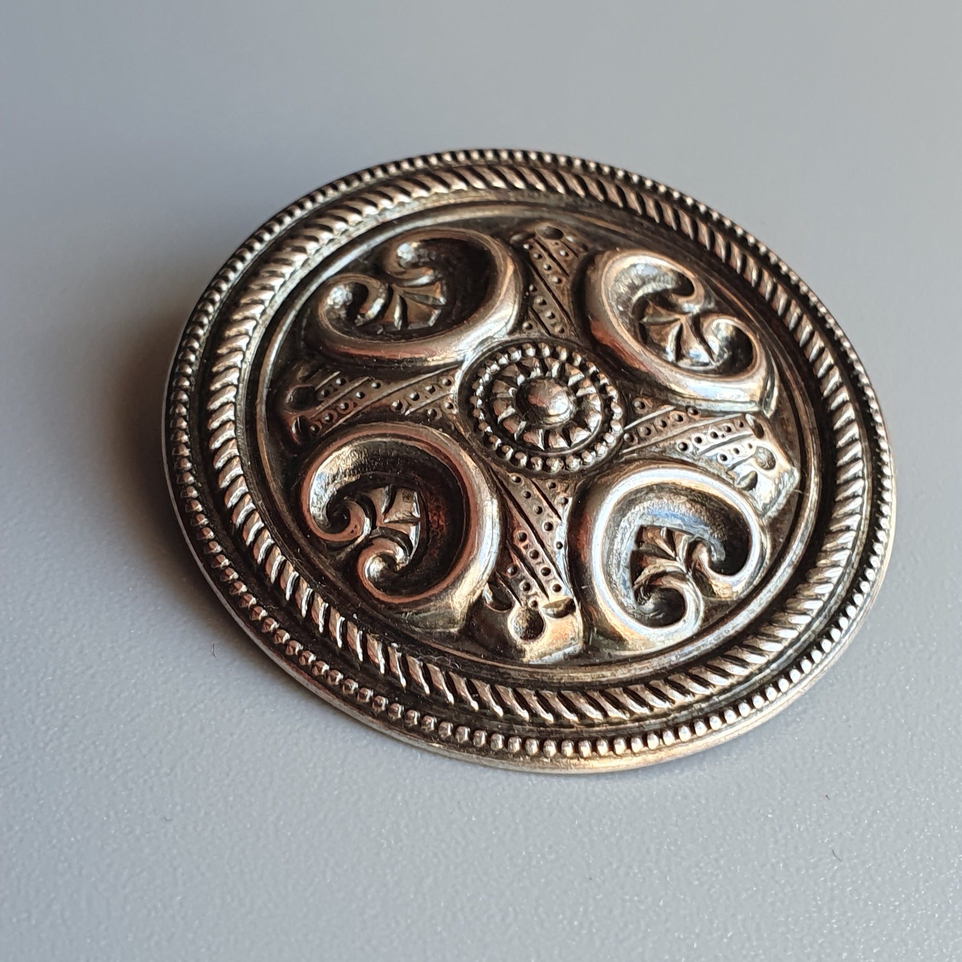 Ornate silver brooch with intricate spiral and floral designs in a circular shape.