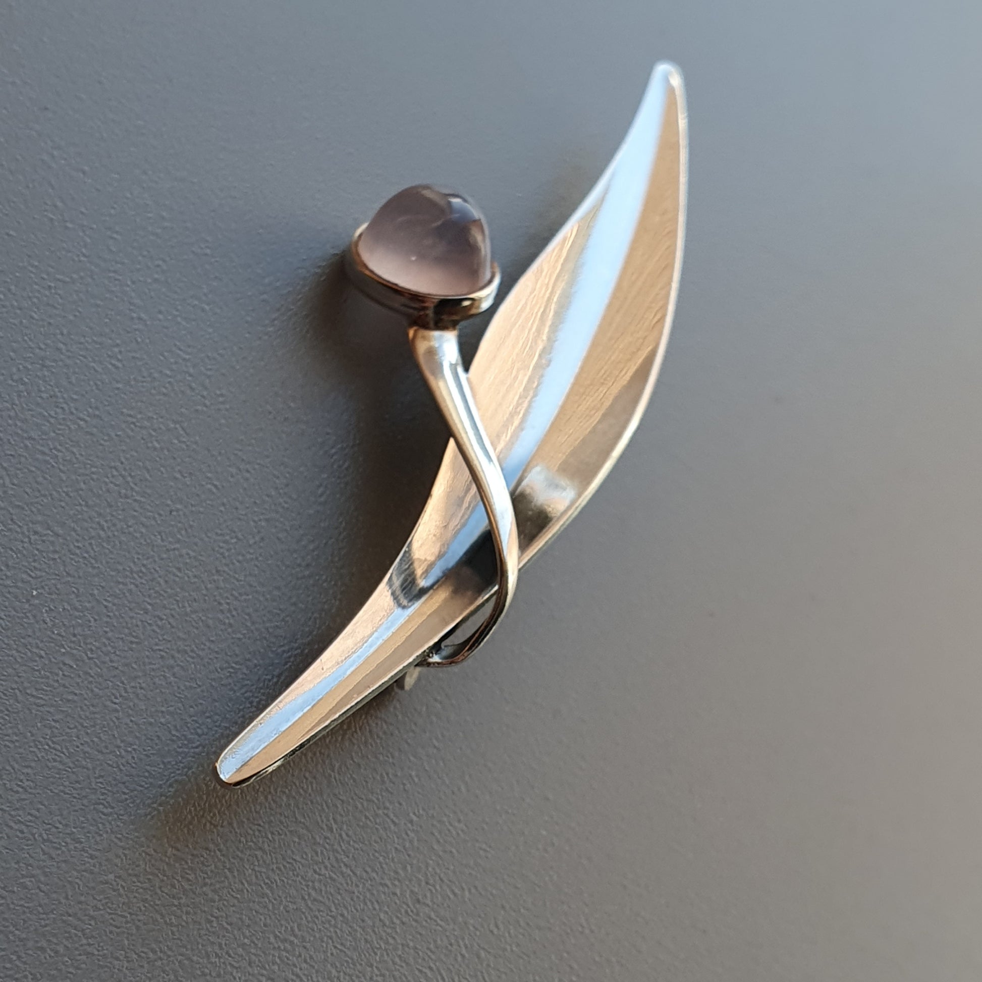 Sleek silver brooch shaped like a stylized leaf or feather with a small gemstone accent.